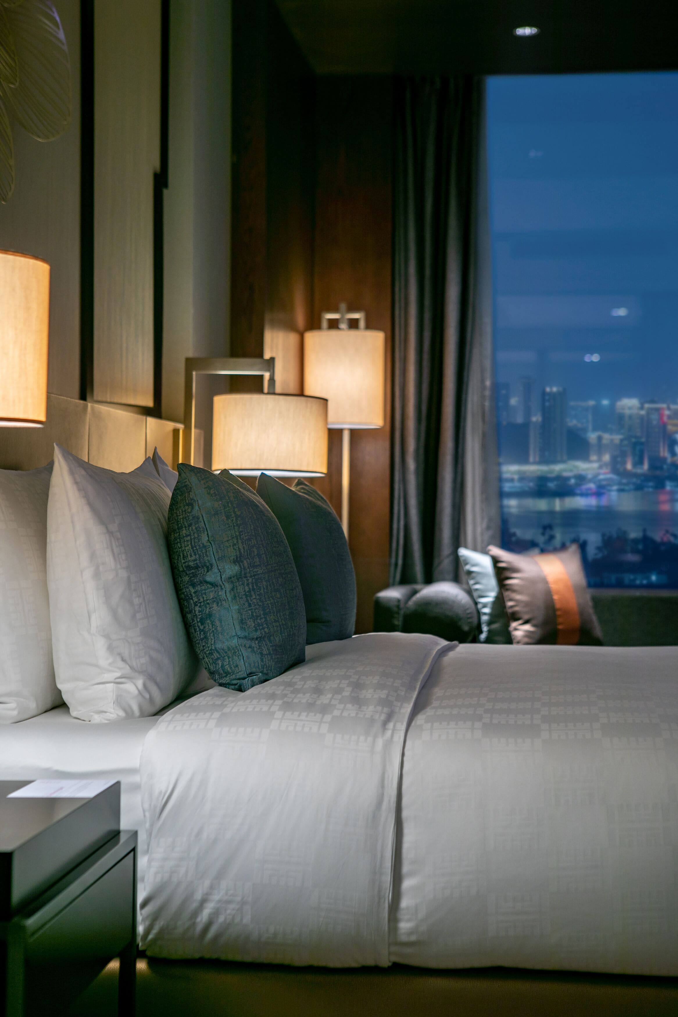 A modern hotel room with a large bed, multiple pillows, bedside lamps, a nightstand, and a view of a cityscape through a window.