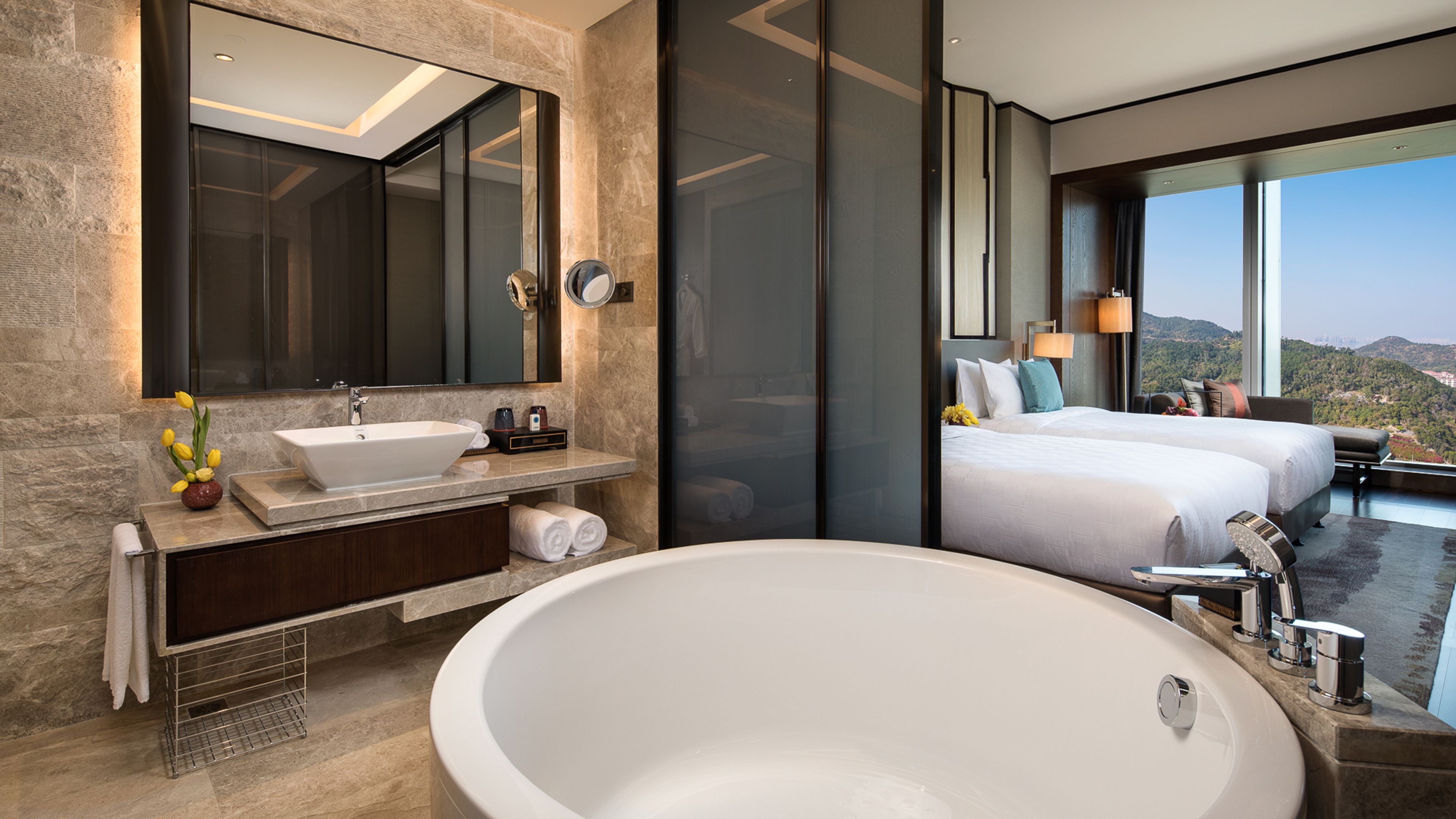 A modern bathroom with a large bathtub, a sink, and a mirror. There is a bed with a view of the mountains in the background.