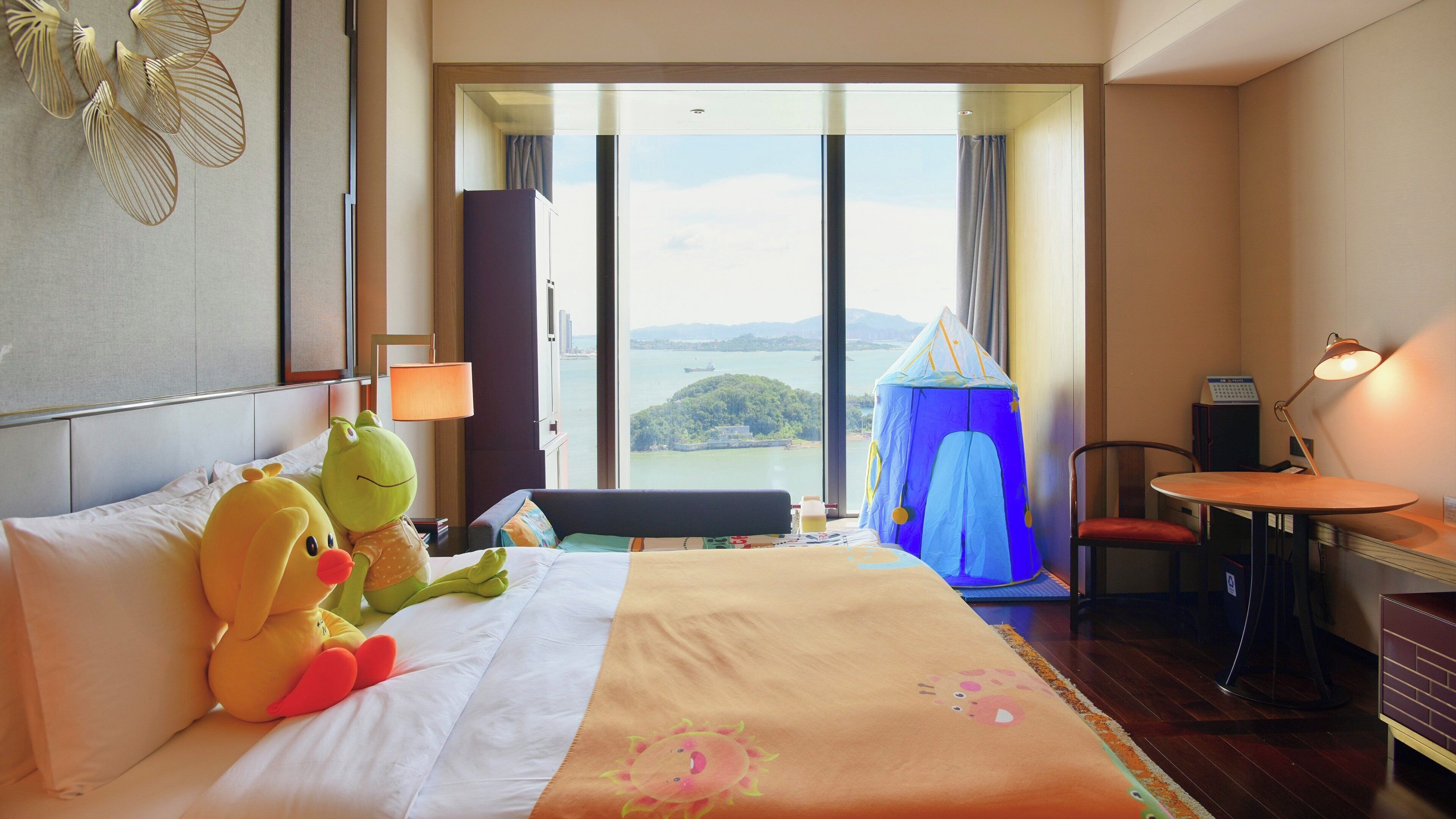 A hotel room with a bed, a small desk, a chair, a lamp, a tent, and a view of the sea.