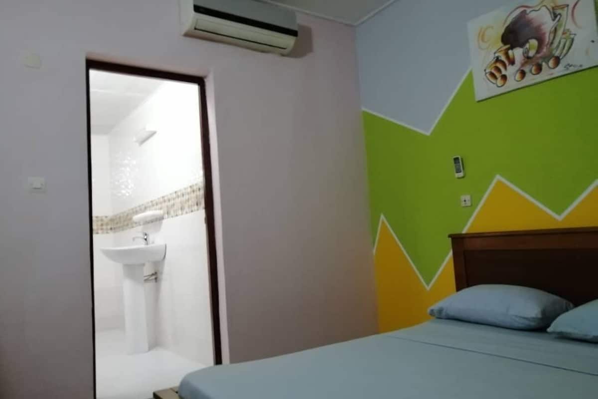 A bedroom with a bed, a wall-mounted air conditioner, a bathroom with a sink, and a colorful wall design.