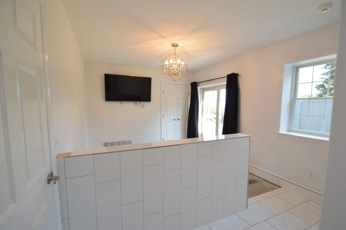 Um quarto com piso de azulejos brancos, porta branca, TV de tela plana presa na parede e janela com vista para o verde lá fora.