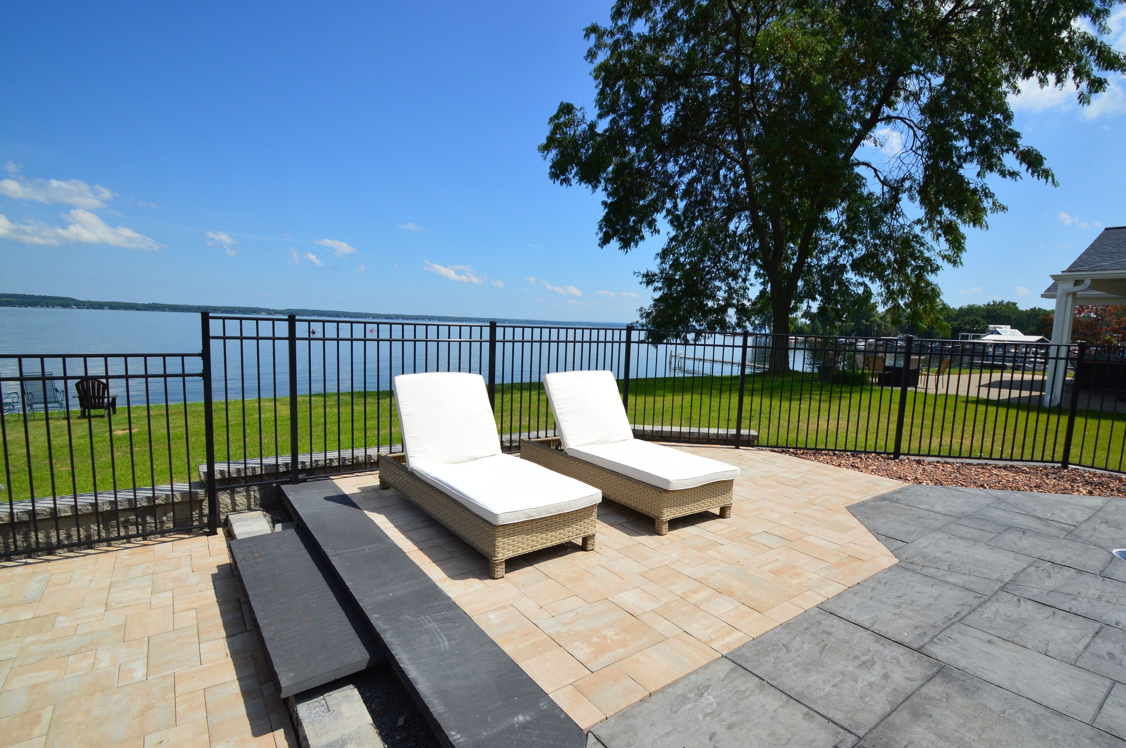 A patio with two lounge chairs and a picnic table, overlooking a body of water and a grassy area.