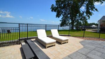 A patio with two lounge chairs and a picnic table, overlooking a body of water and a grassy area.