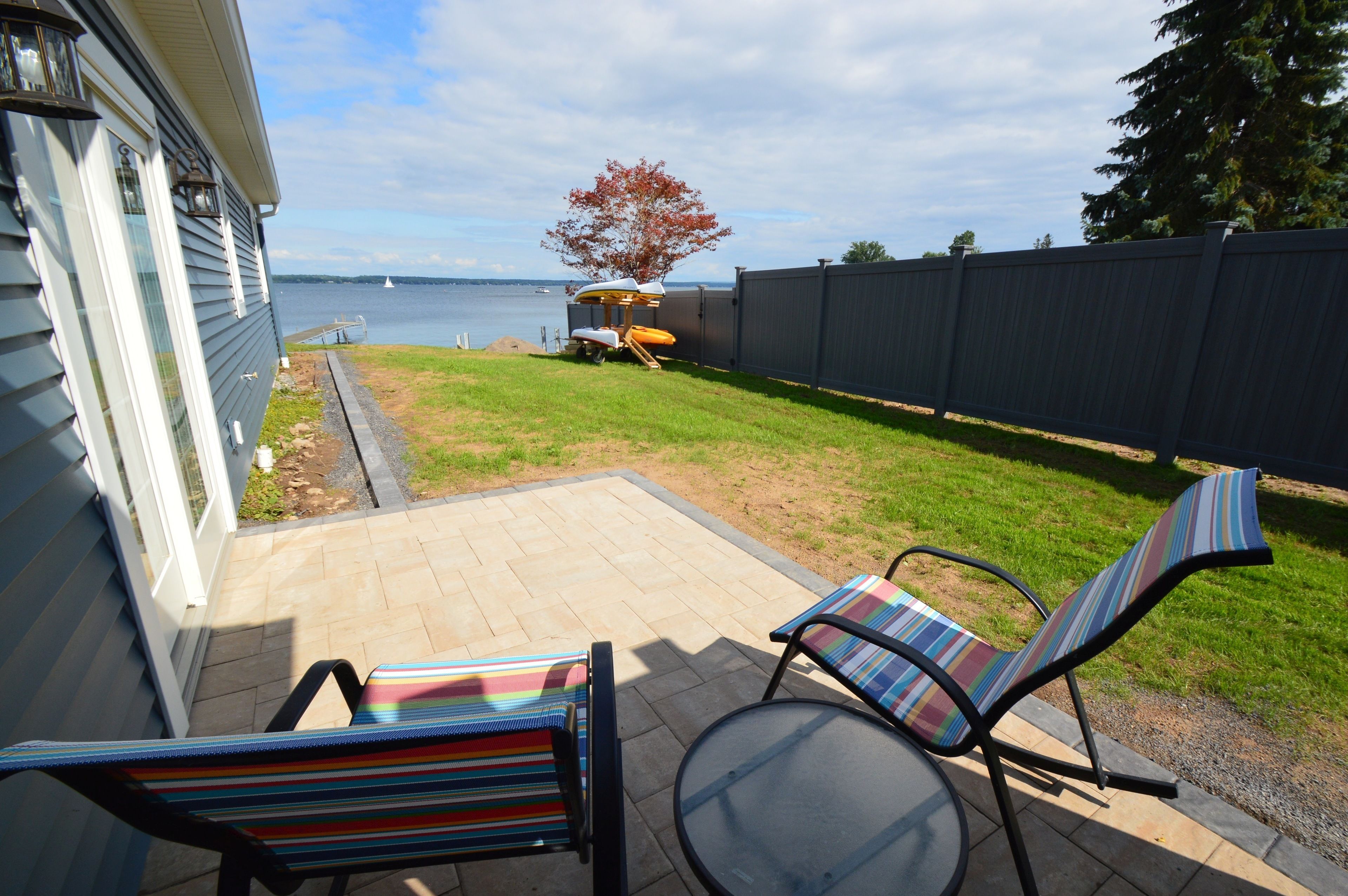 A patio with two striped chairs and a table, a grassy yard, a wooden fence, and a view of a body of water.