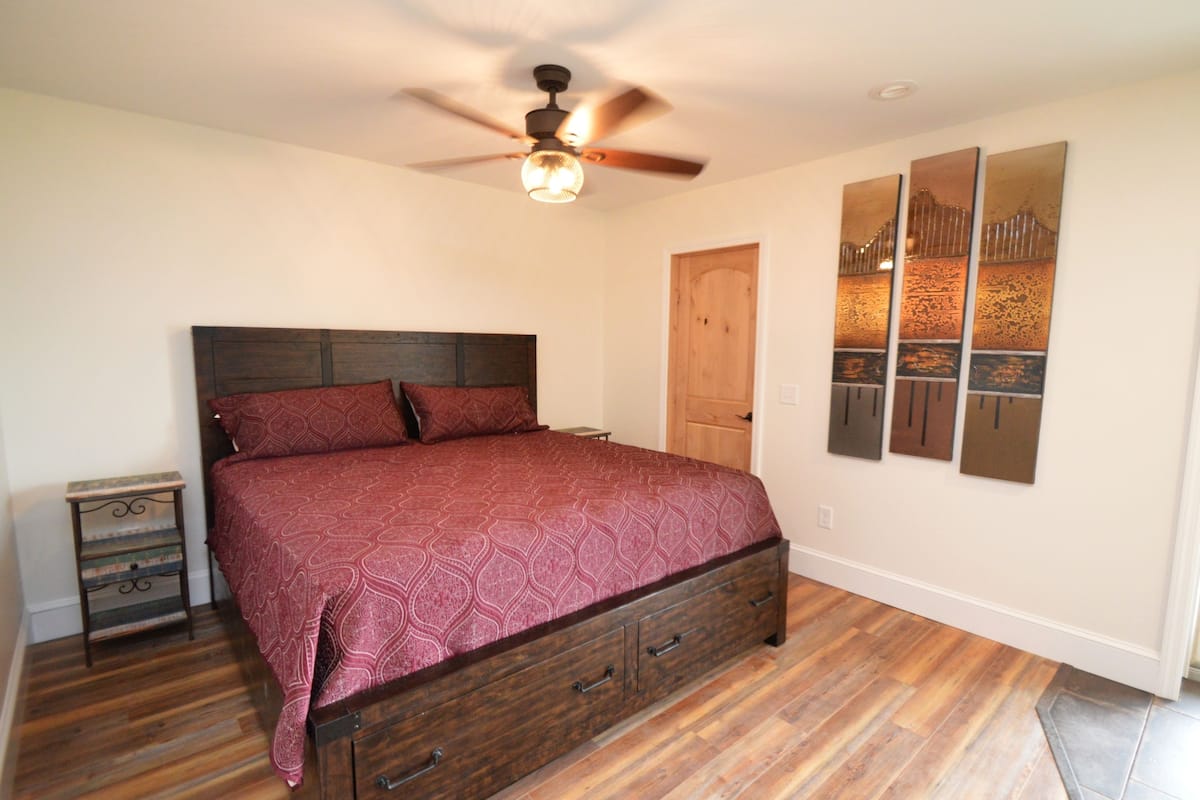 Quarto com cama de madeira, mesa de cabeceira, ventilador de teto e porta.