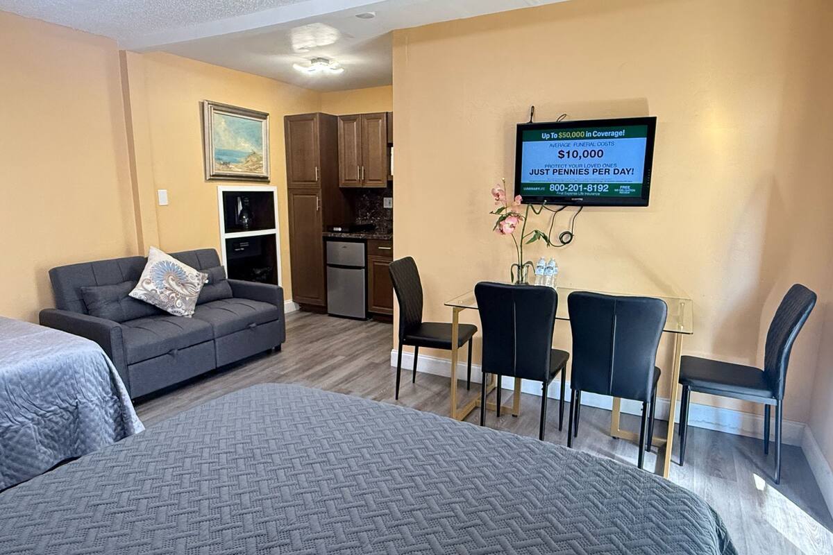 A hotel room with a bed, a sofa, a dining table with chairs, a kitchen area, and a TV displaying a message about funeral costs.
