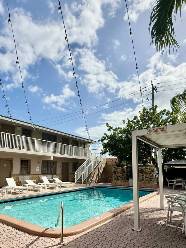 A pool area with lounge chairs, a covered seating area, and a building with a balcony.