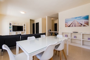 Un salon moderne avec une table à manger blanche, un canapé noir et un téléviseur posé sur une étagère blanche.