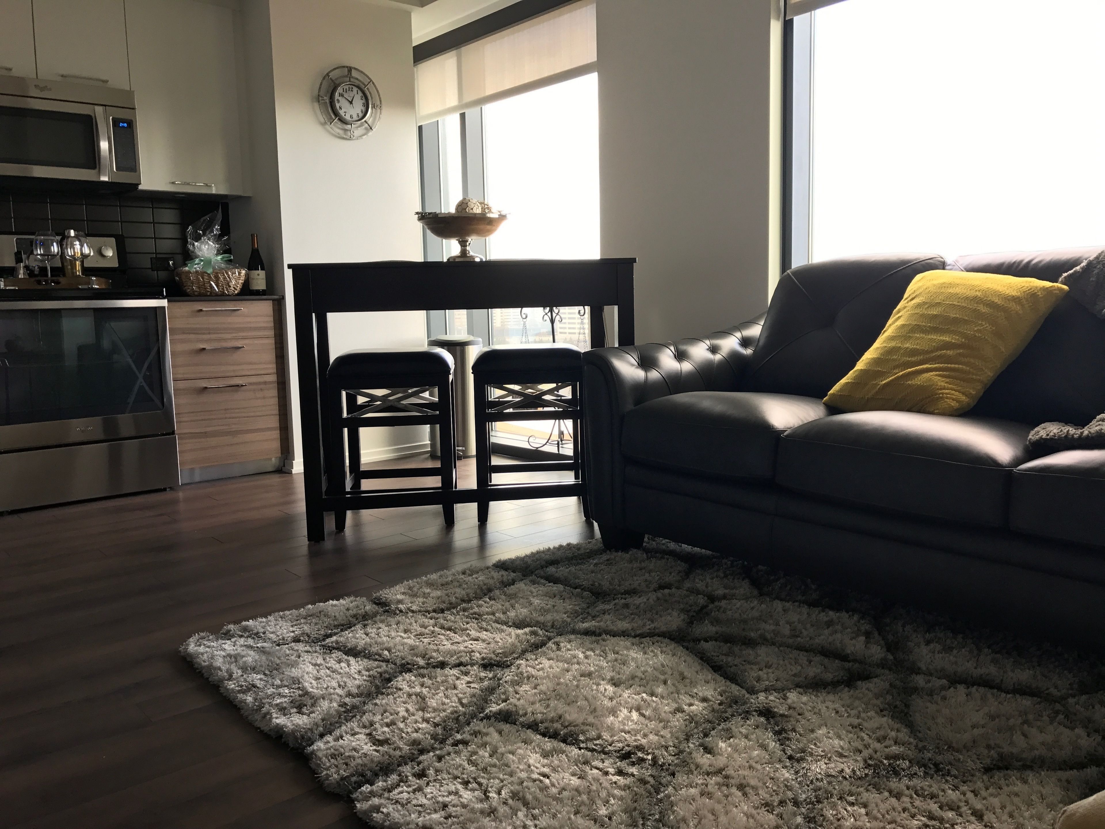A modern kitchen with stainless steel appliances, a dining area with a black table and two chairs, and a living room with a dark leather sofa and a fluffy rug.