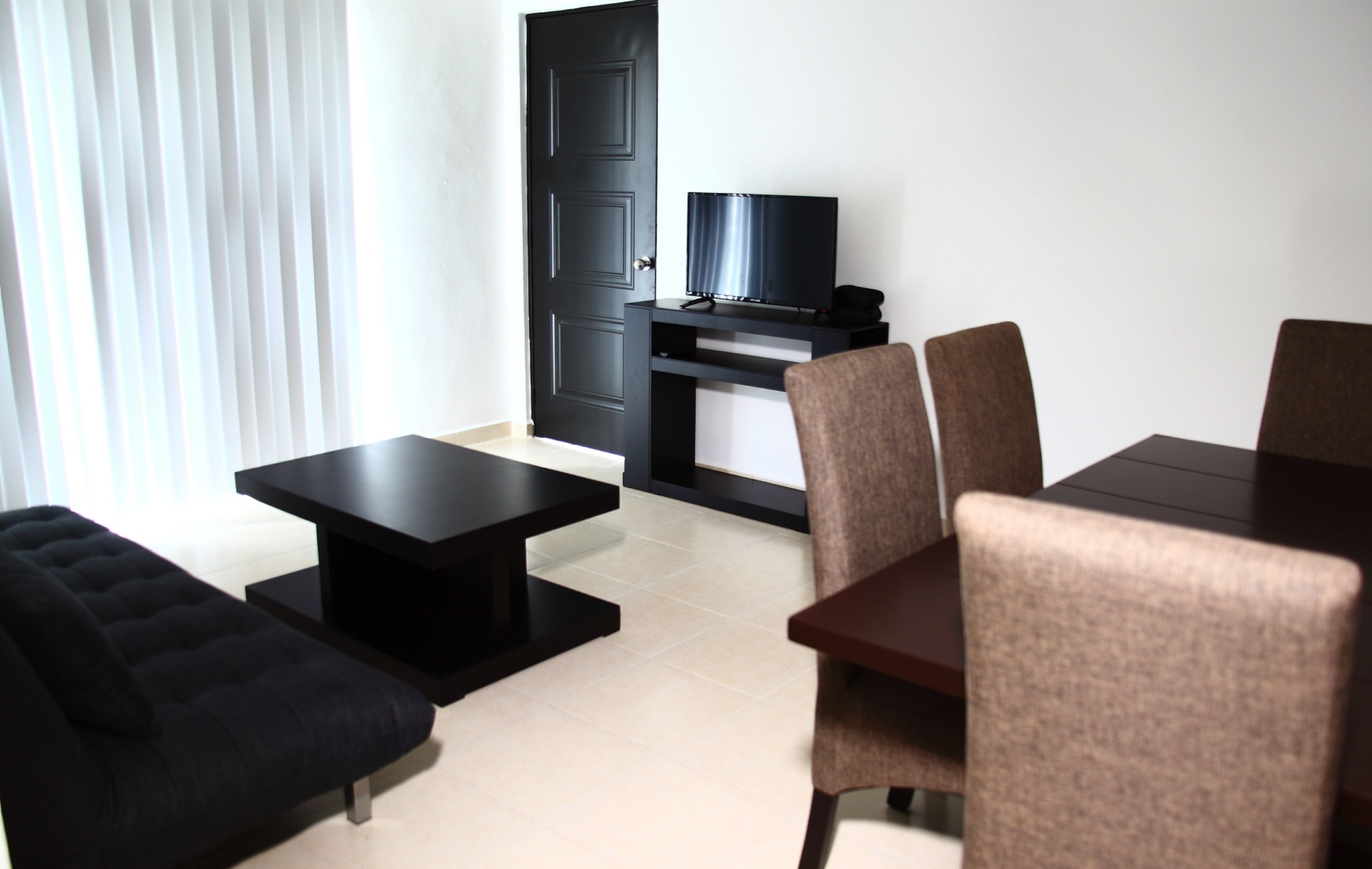 Una sala de estar moderna con un sofá negro, una mesa de centro, un televisor sobre un mueble bajo y una mesa de comedor con sillas.