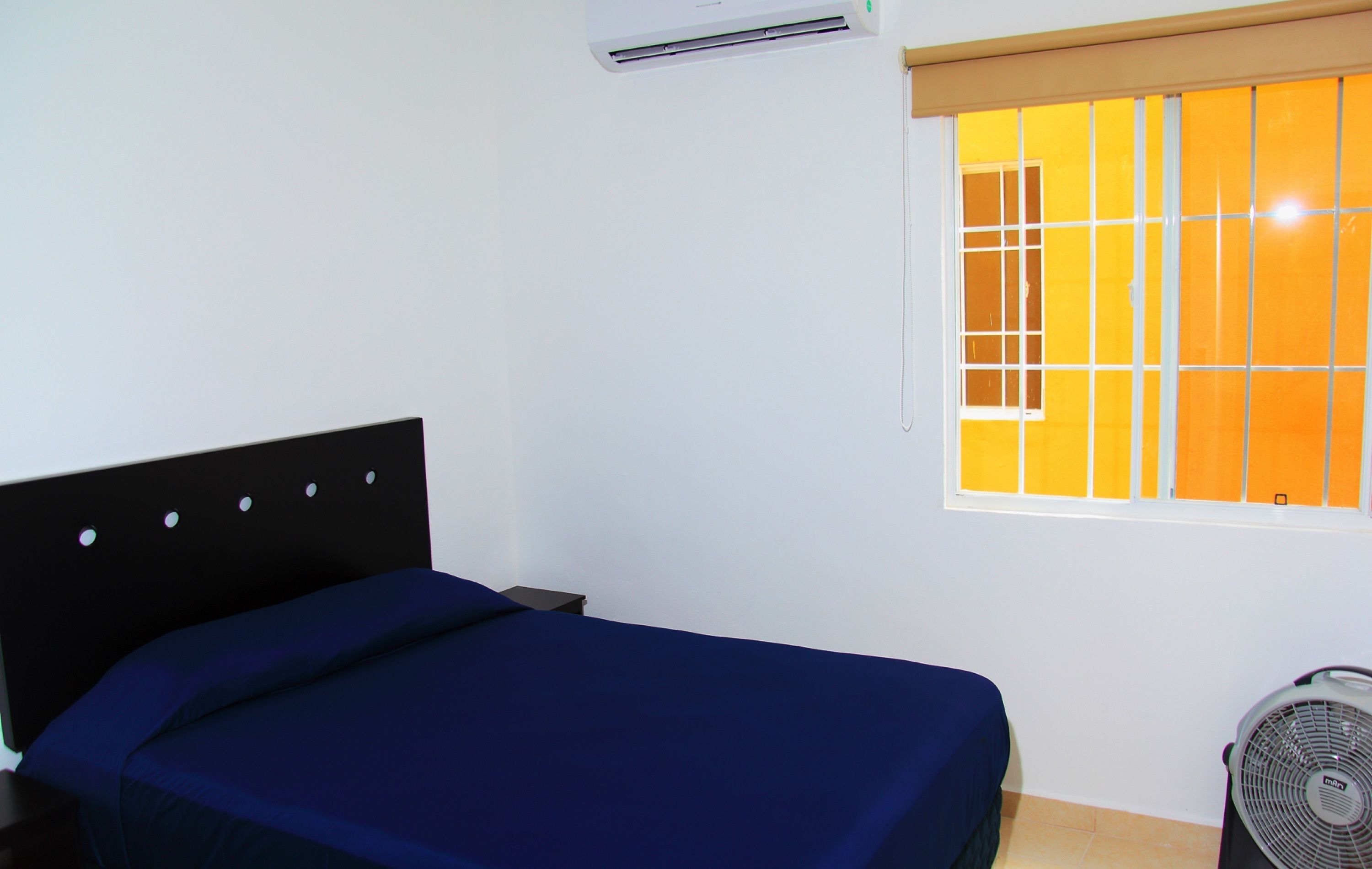 Une chambre avec un lit, une fenêtre avec des rideaux orange, un climatiseur et un ventilateur sur pied.