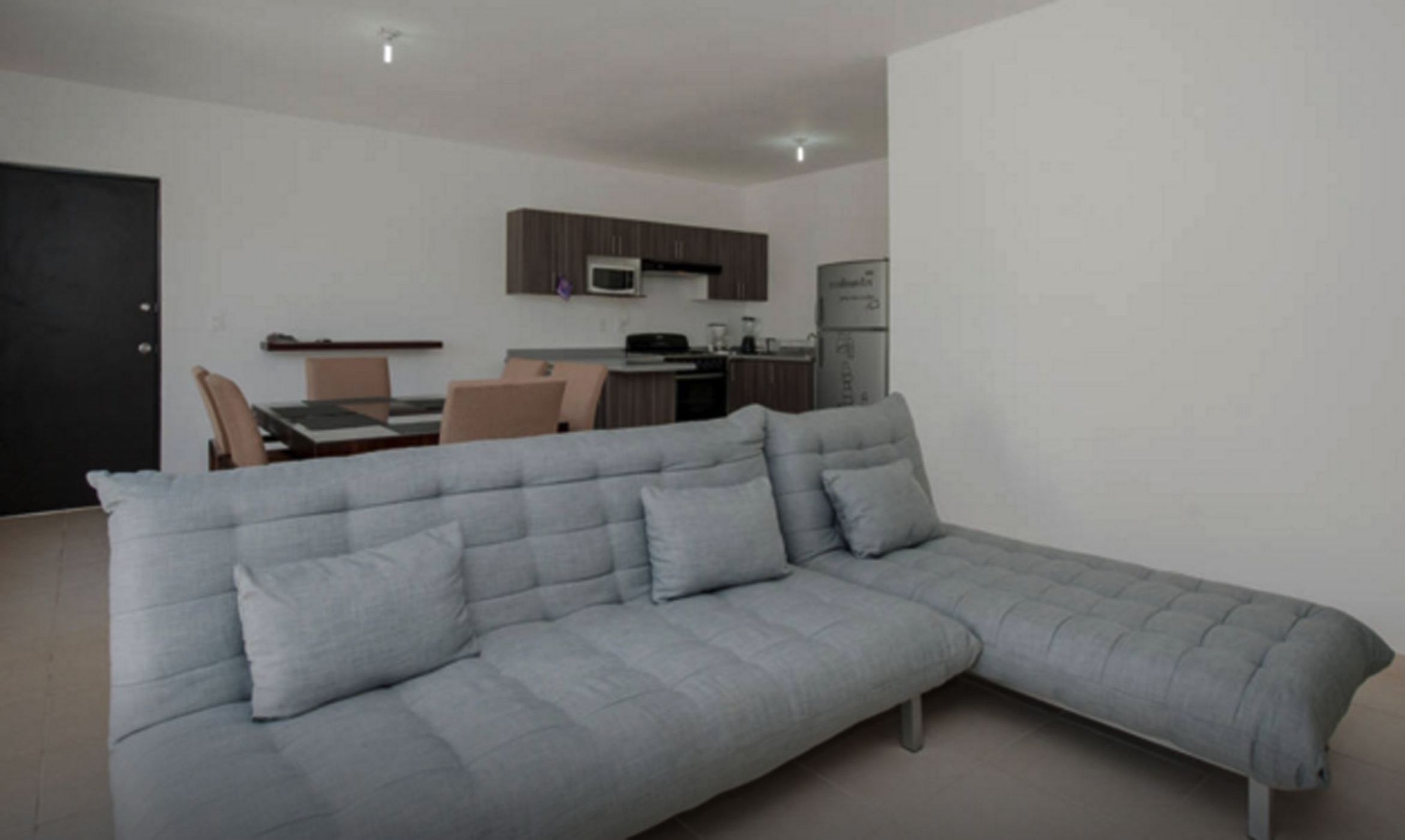 A modern living room with a grey sectional sofa, a dining area with a table and chairs, and a kitchen with wooden cabinets and appliances.