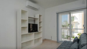 A modern living room with a grey sofa, a flat-screen TV on a white shelving unit, and a sliding glass door leading to a balcony. - DEPARTAMENTO VELAS SELVANOVA 512 (Playa del Carmen)