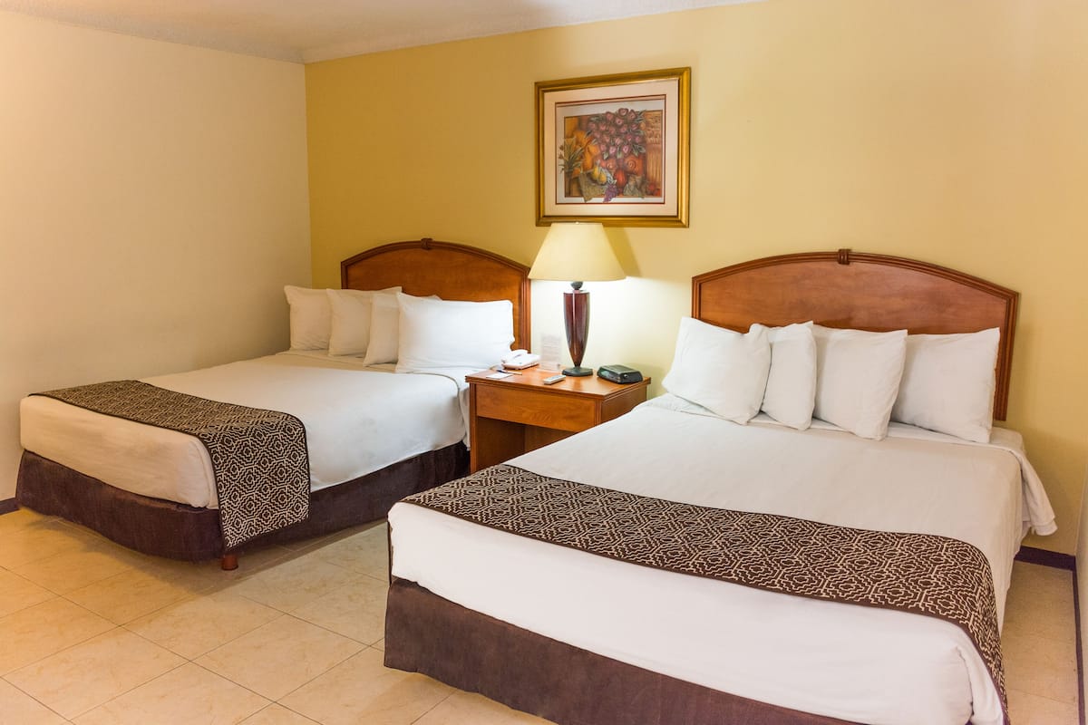 A hotel room with two beds, each with a wooden headboard, a bedside table with a lamp, and a framed artwork on the wall.