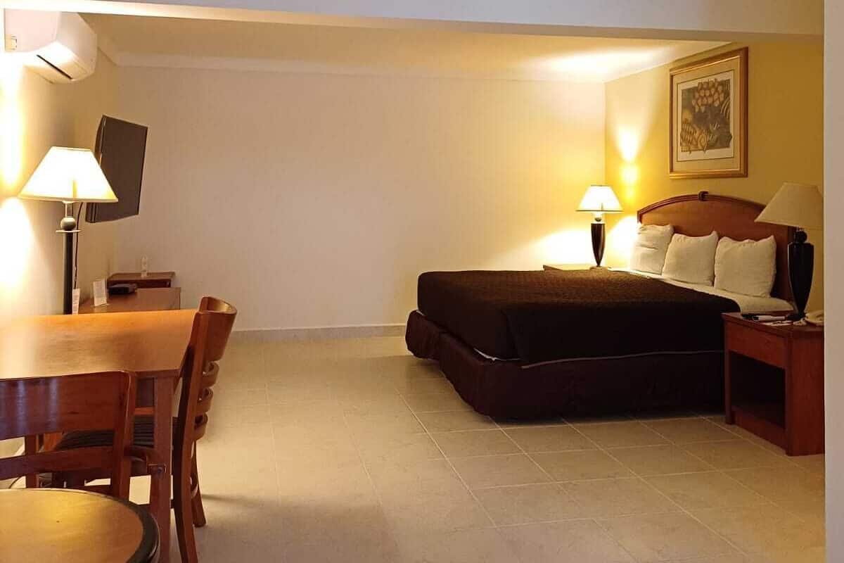 A compact hotel room with a bed, a small dining table, and chairs. There is a TV mounted on the wall and a framed picture above the bed.