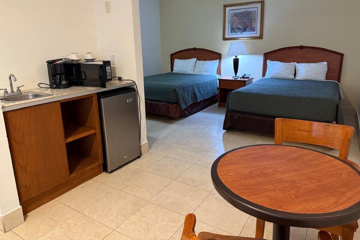 A compact hotel room with a kitchenette, two single beds, a small dining table, and a chair.