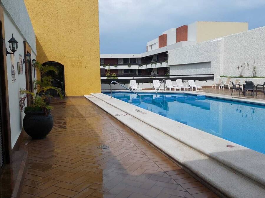 A swimming pool with lounge chairs, a potted plant, and a building with balconies in the background.