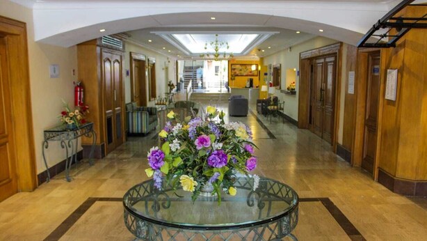 A hotel lobby with a central glass table surrounded by a floral arrangement, wooden doors, and a staircase in the background.