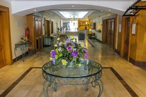 A hotel lobby with a central glass table surrounded by a floral arrangement, wooden doors, and a staircase in the background.