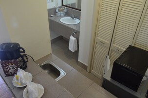 Un baño pequeño con lavamanos, espejo y barra para toallas. Hay una cafetera y un cubo de basura en la encimera.
