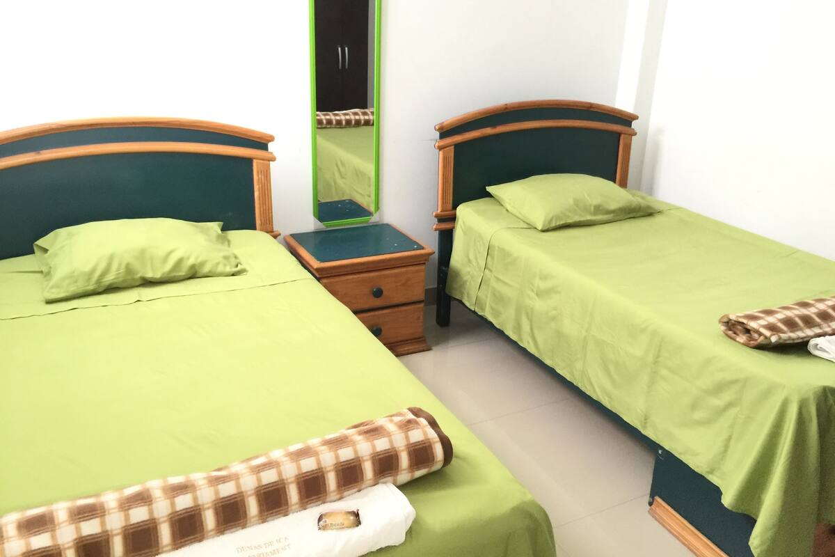 Una habitación con dos camas individuales, cada una con una colcha verde y un cojín a cuadros marrón. Hay una mesita de noche de madera con un espejo entre las camas.