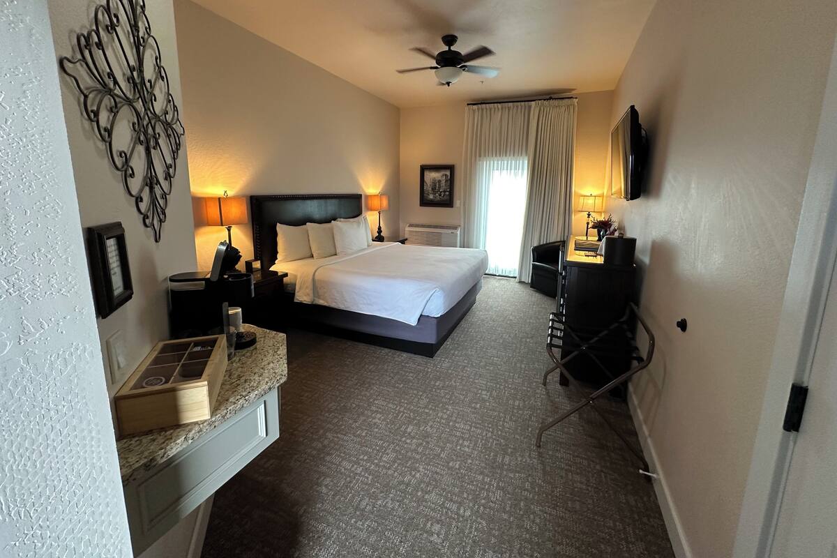 A hotel room with a large bed, a desk with a chair, a vanity with a mirror, and a ceiling fan.