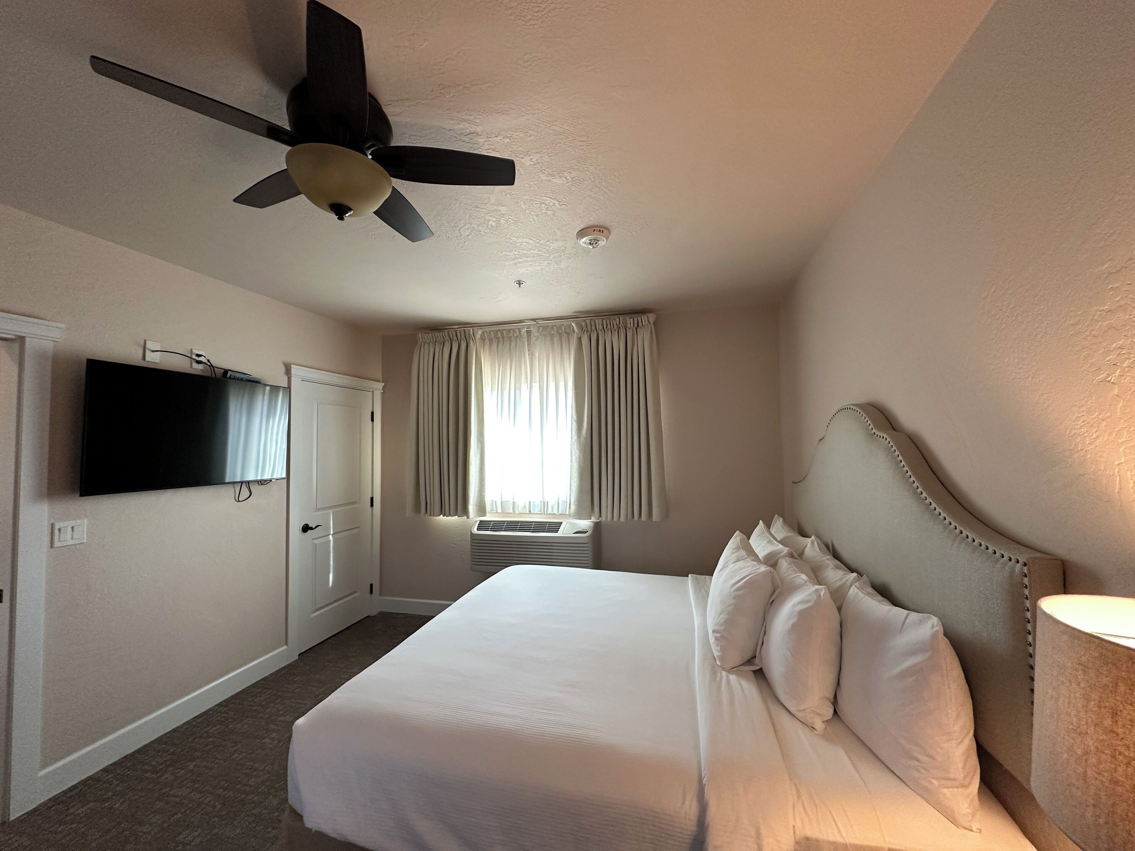 A hotel room with a bed, a ceiling fan, a TV, a window with curtains, and an air conditioning unit.