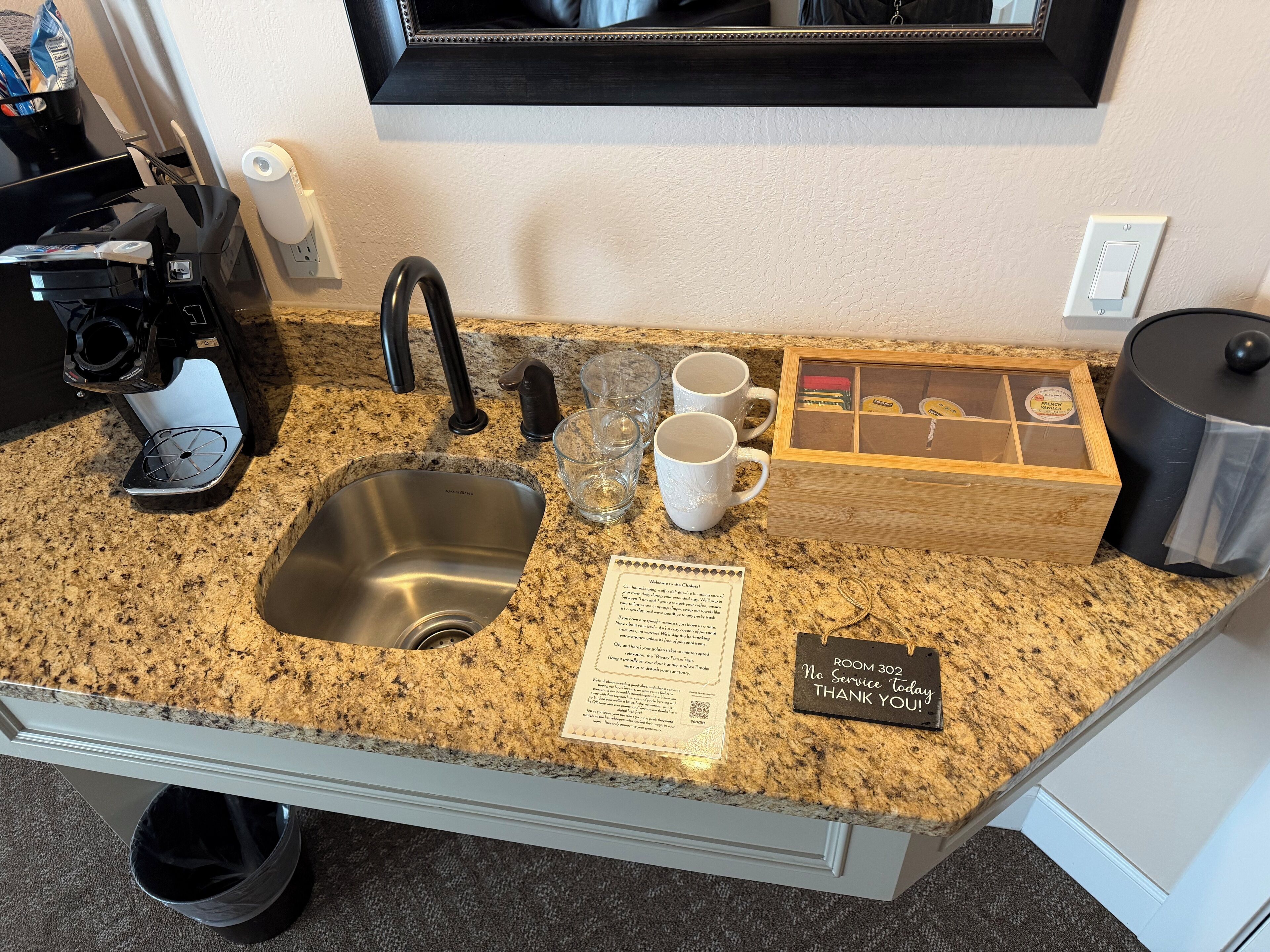 A kitchen counter with a sink, coffee maker, mugs, and a tea box.