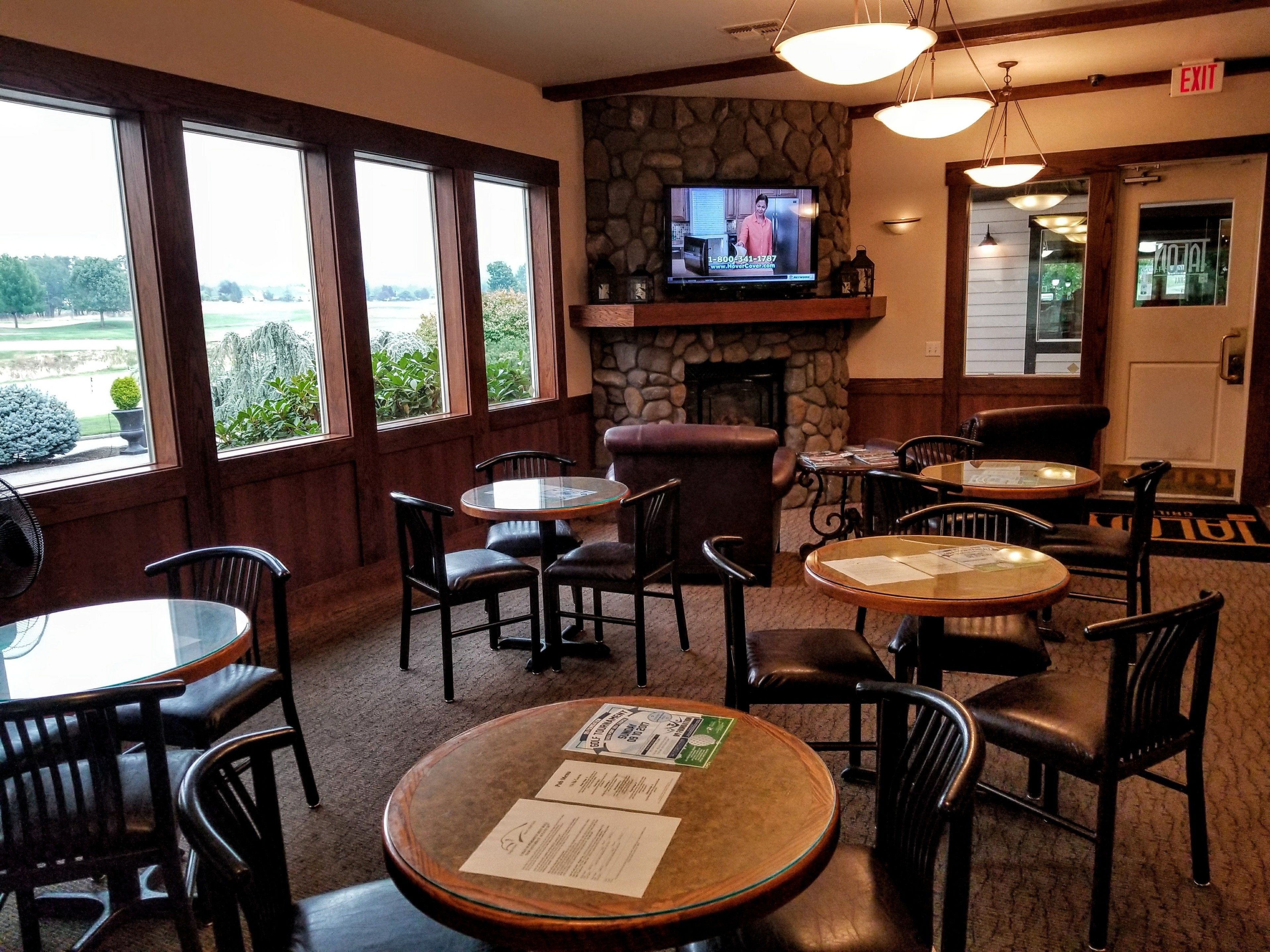 A restaurant with multiple tables and chairs, a stone fireplace, a television mounted on the wall, and large windows offering a view of the outdoors.