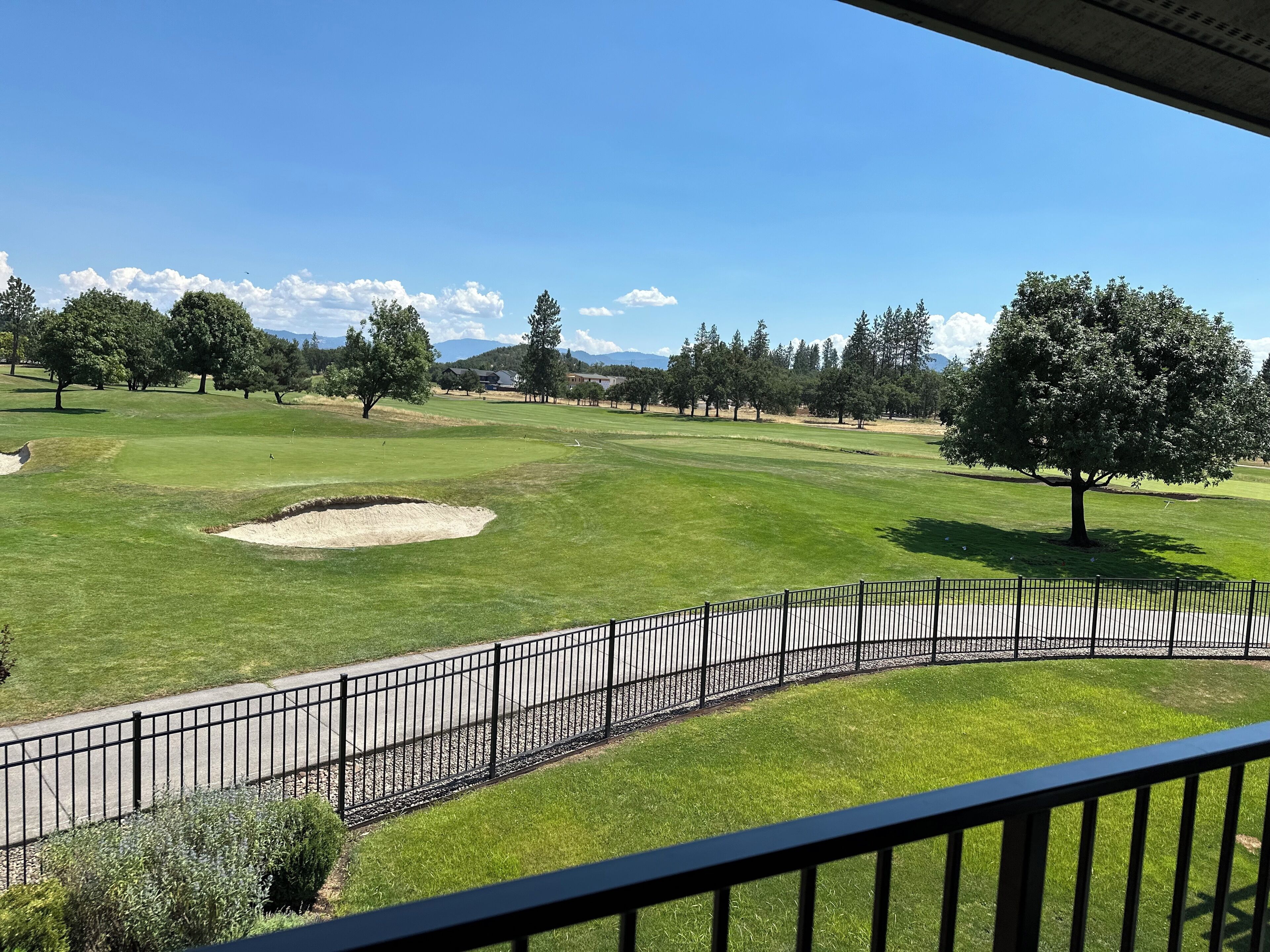 A view of a golf course with a clear sky, a fenced area, and a large tree in the foreground.