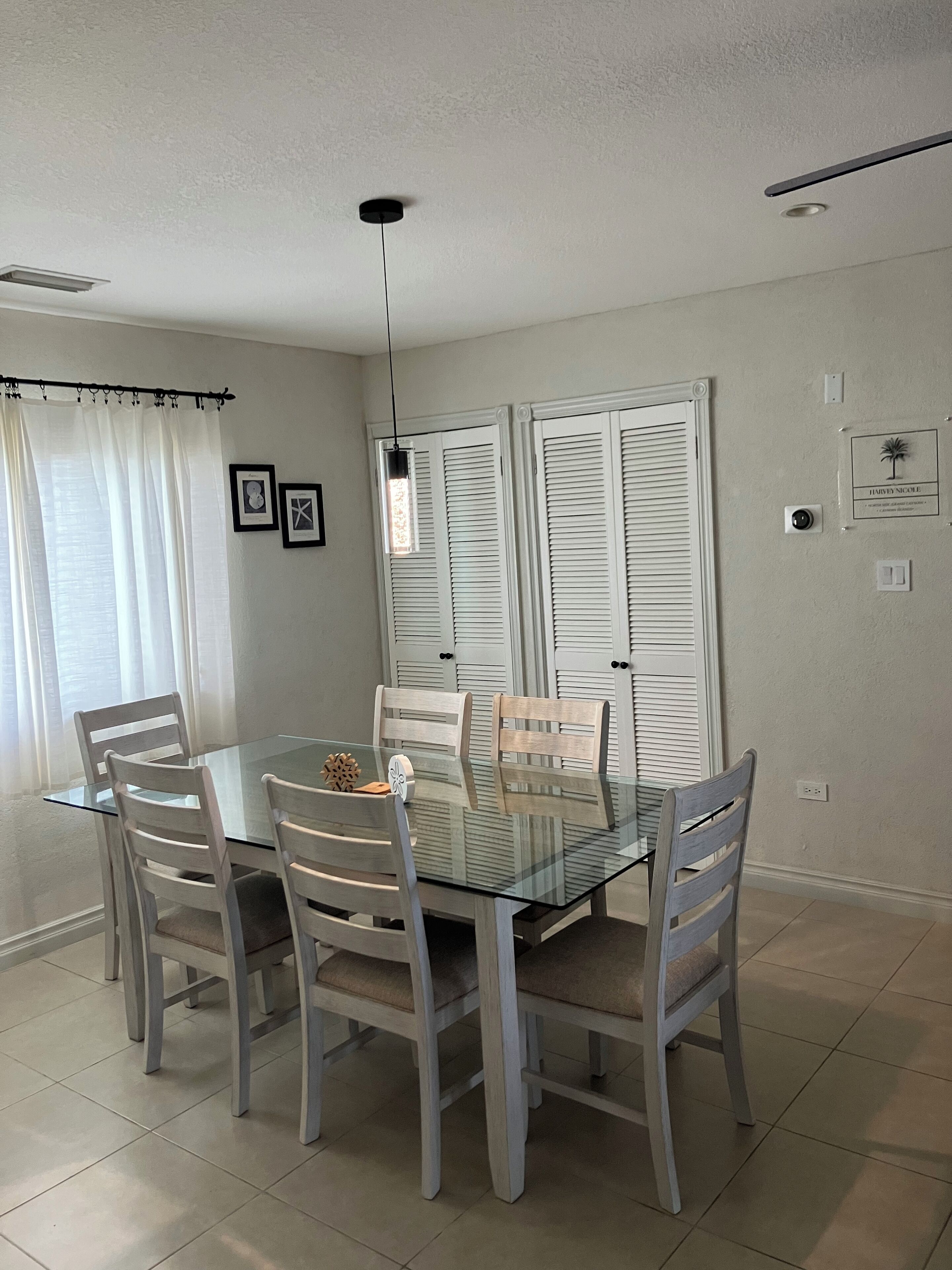 A dining area with a glass table surrounded by wooden chairs, white shutters on the windows, and a ceiling light fixture.