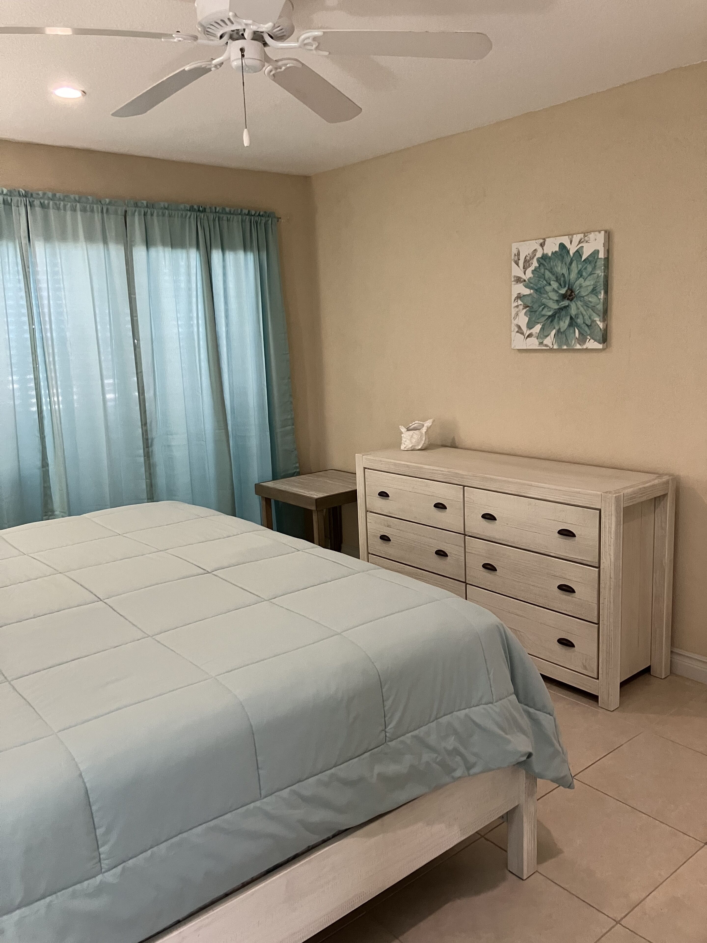 Un dormitorio con cama, cómoda, ventilador de techo y ventana con cortinas.