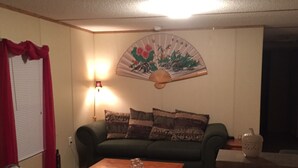 Una sala de estar con un sofá verde, una mesa de centro de madera, un ventilador, una lámpara y un abanico decorativo en la pared.