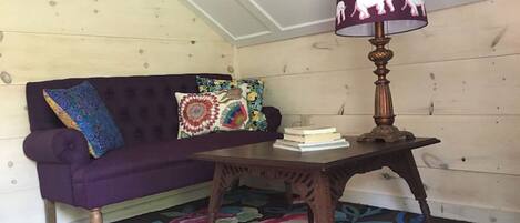 Une pièce chaleureuse avec un canapé violet, des coussins aux couleurs vives, une table en bois surmontée d’une lampe et un tapis éclatant.