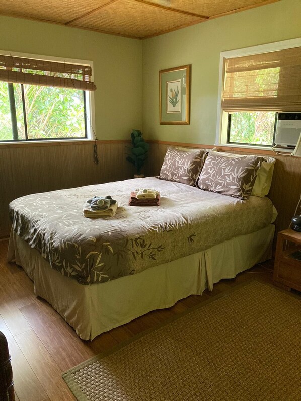 A neatly made bed with a floral bedspread, a bedside table with a lamp, and a window with a view of greenery.
