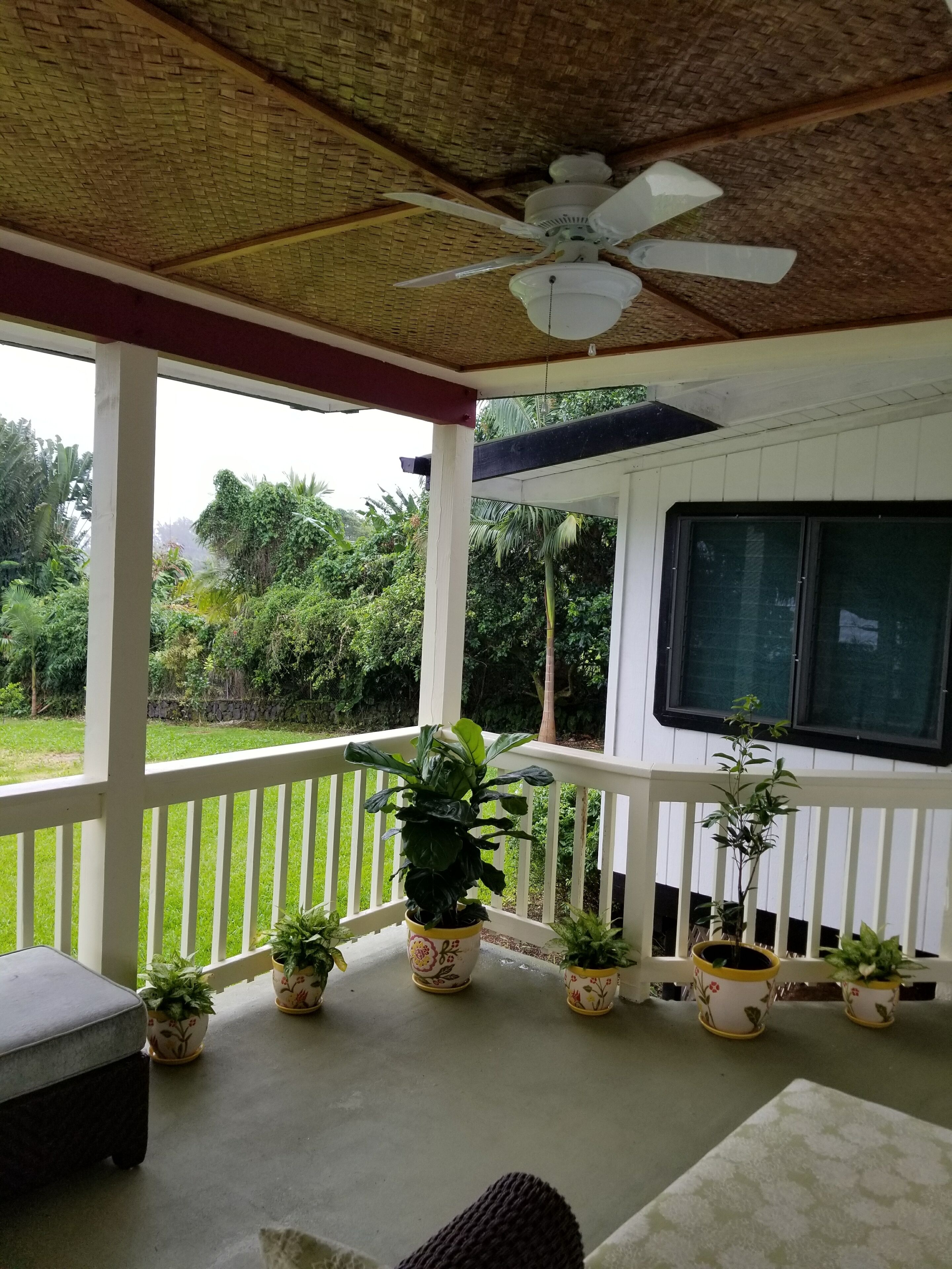 A covered porch with a ceiling fan, potted plants, a bench, and a window overlooking a garden.