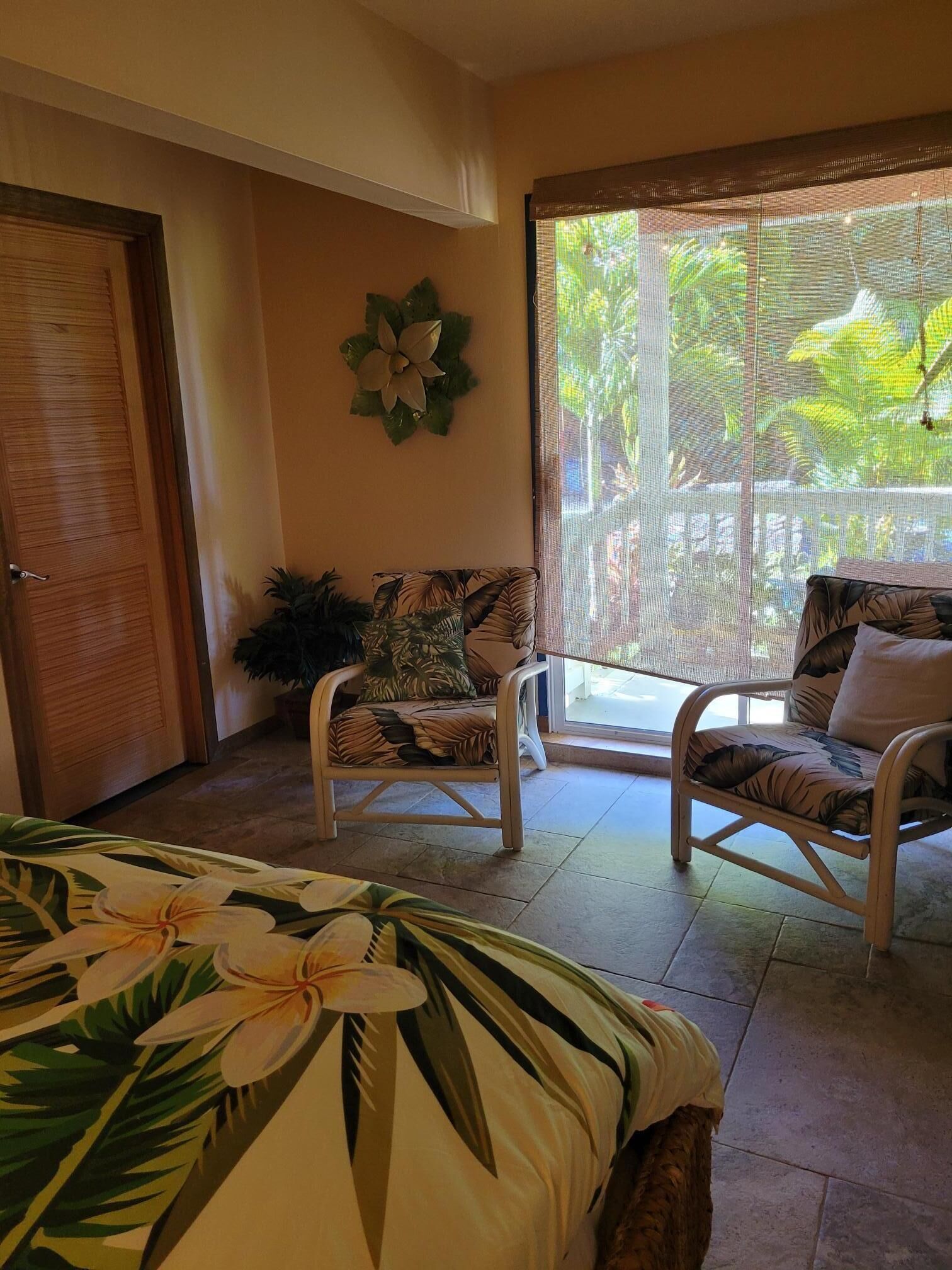 A room with a bed, two wicker chairs, a small table, a decorative wall piece, and a large window with a view of greenery.