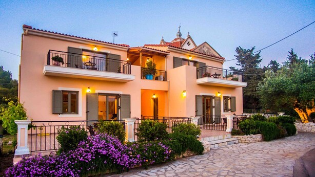 Une maison à deux étages avec un balcon, un chemin de pierre et des fleurs violettes au premier plan.