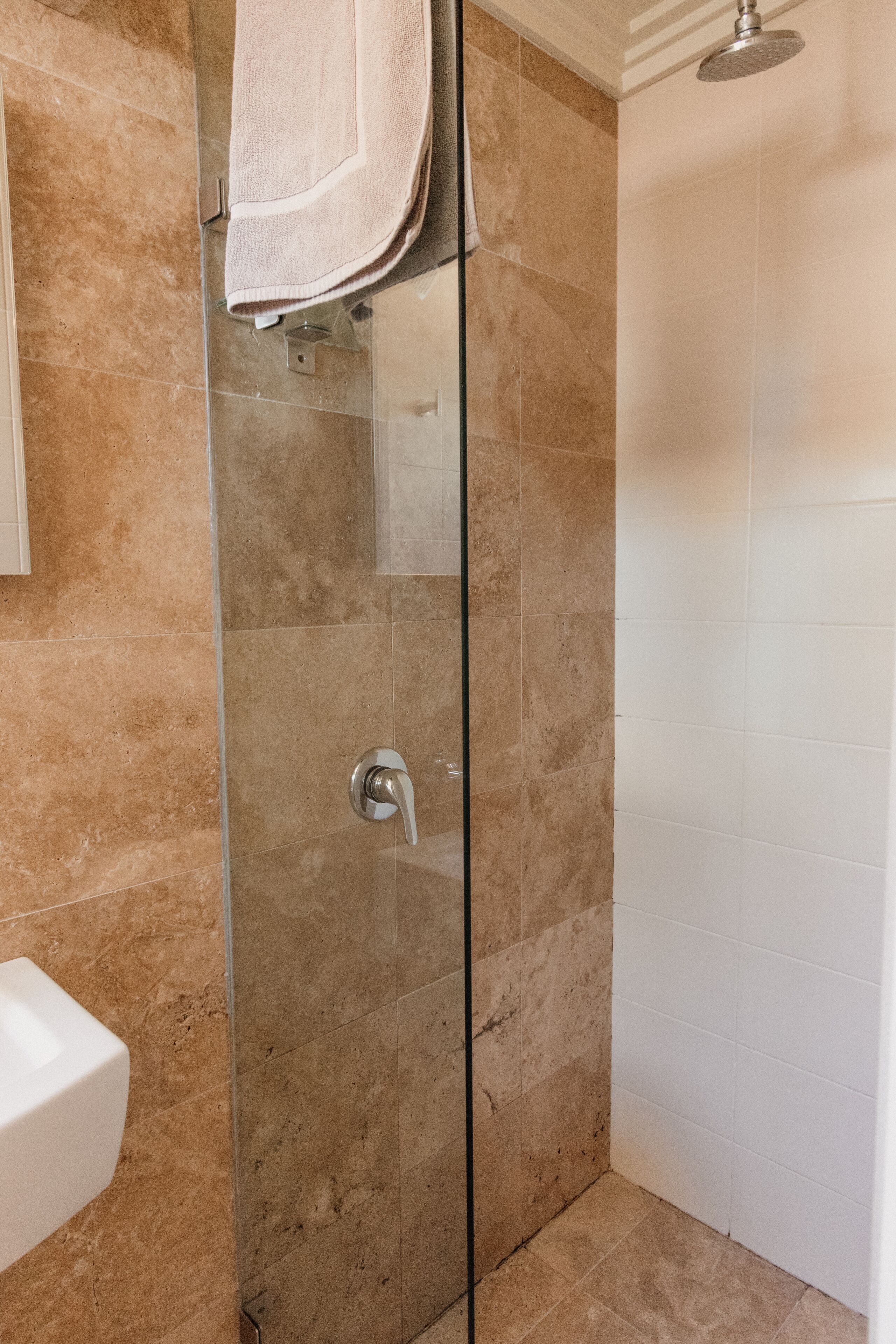 Une douche fermée par des parois de verre, avec des carreaux de couleur beige, un lavabo blanc et une serviette accrochée au mur.