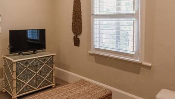 Una sala de estar con televisión sobre un mueble bajo, un banco de mimbre y una ventana con persianas.