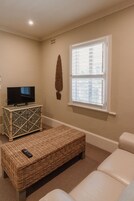 Una sala de estar con televisión sobre un mueble bajo, un banco de mimbre y una ventana con persianas.