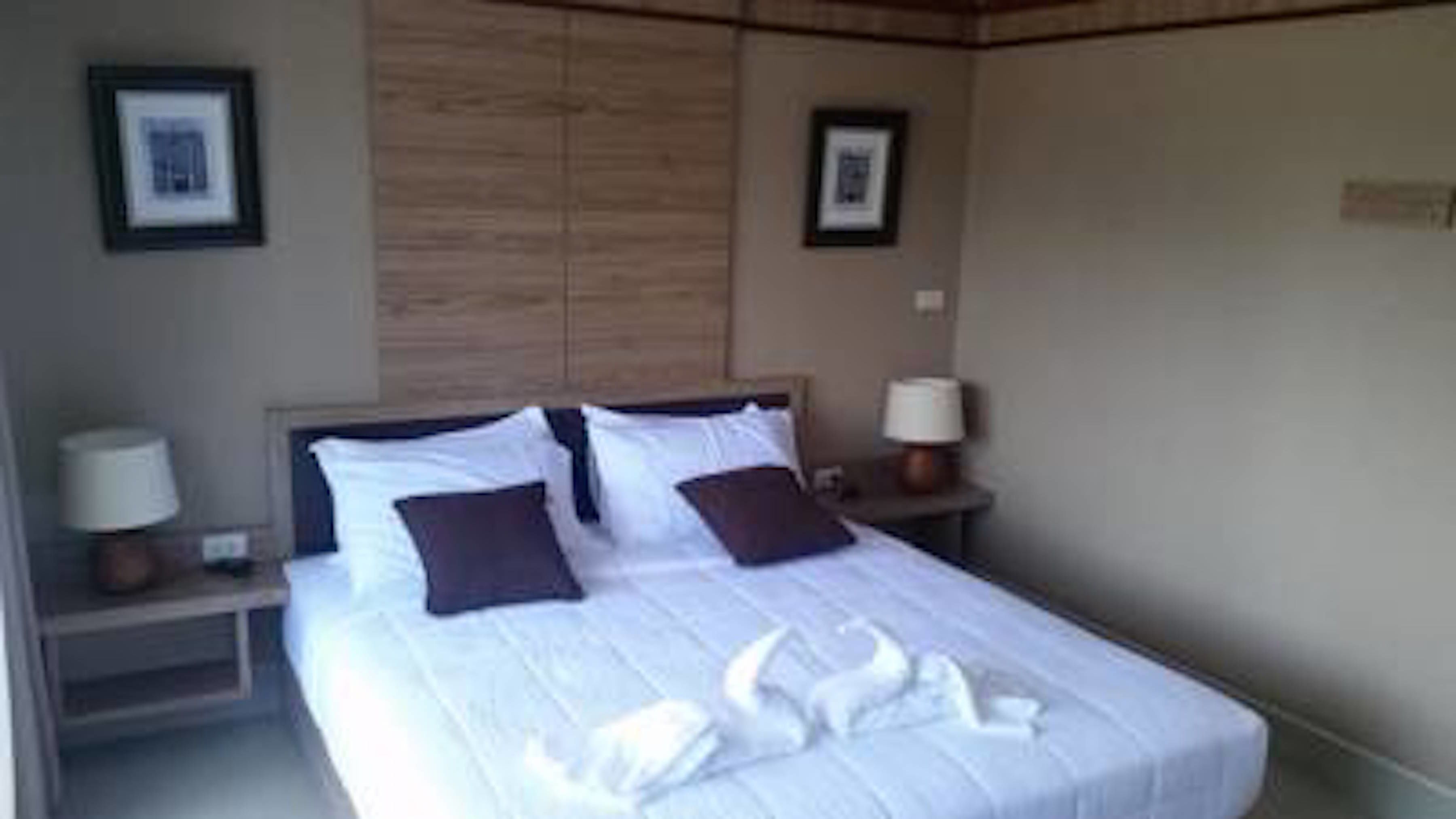 A neatly made bed with white linens and two dark pillows, flanked by two bedside tables with lamps, in a room with a wooden headboard and framed pictures on the walls.