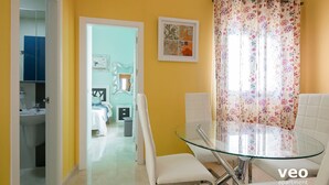 Une pièce lumineuse aux murs jaune vif, avec une table à manger en verre, des chaises blanches et un rideau à motifs floraux.