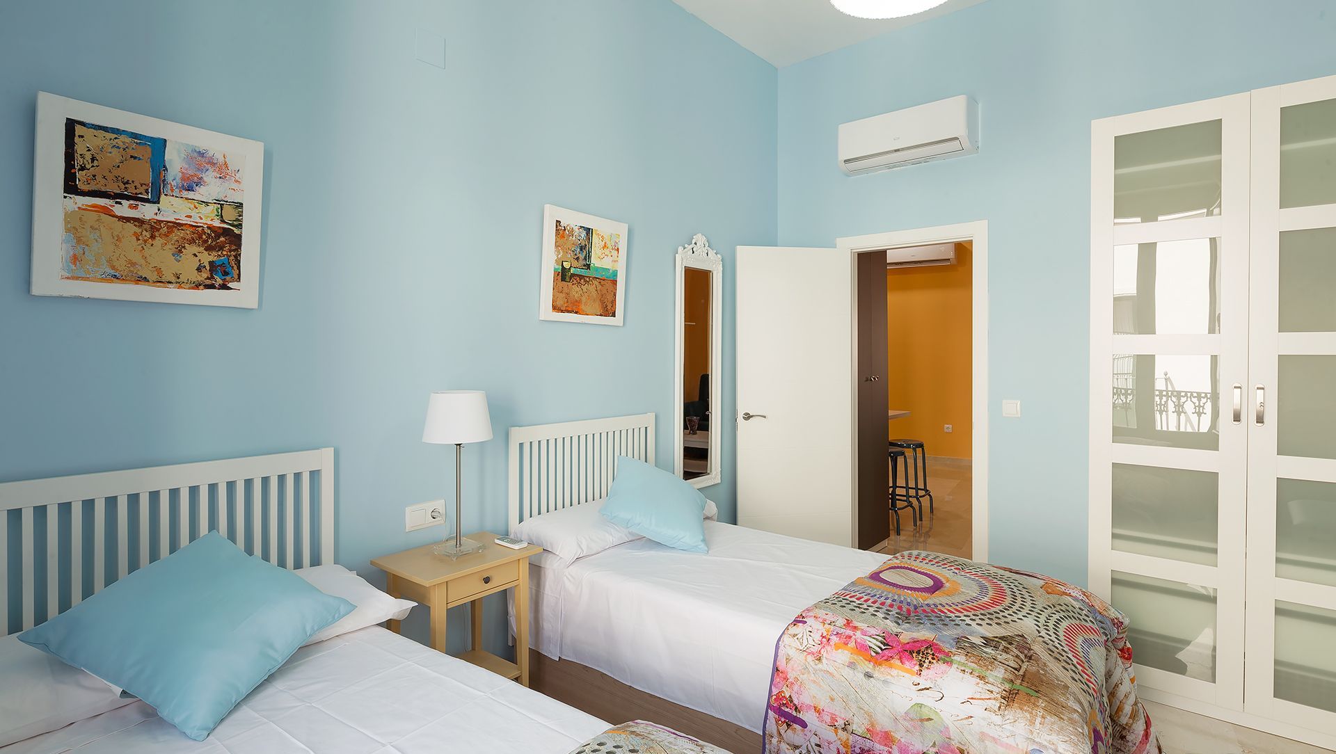 Quarto com duas camas, uma mesa de cabeceira, uma luminária e uma porta para o banheiro.