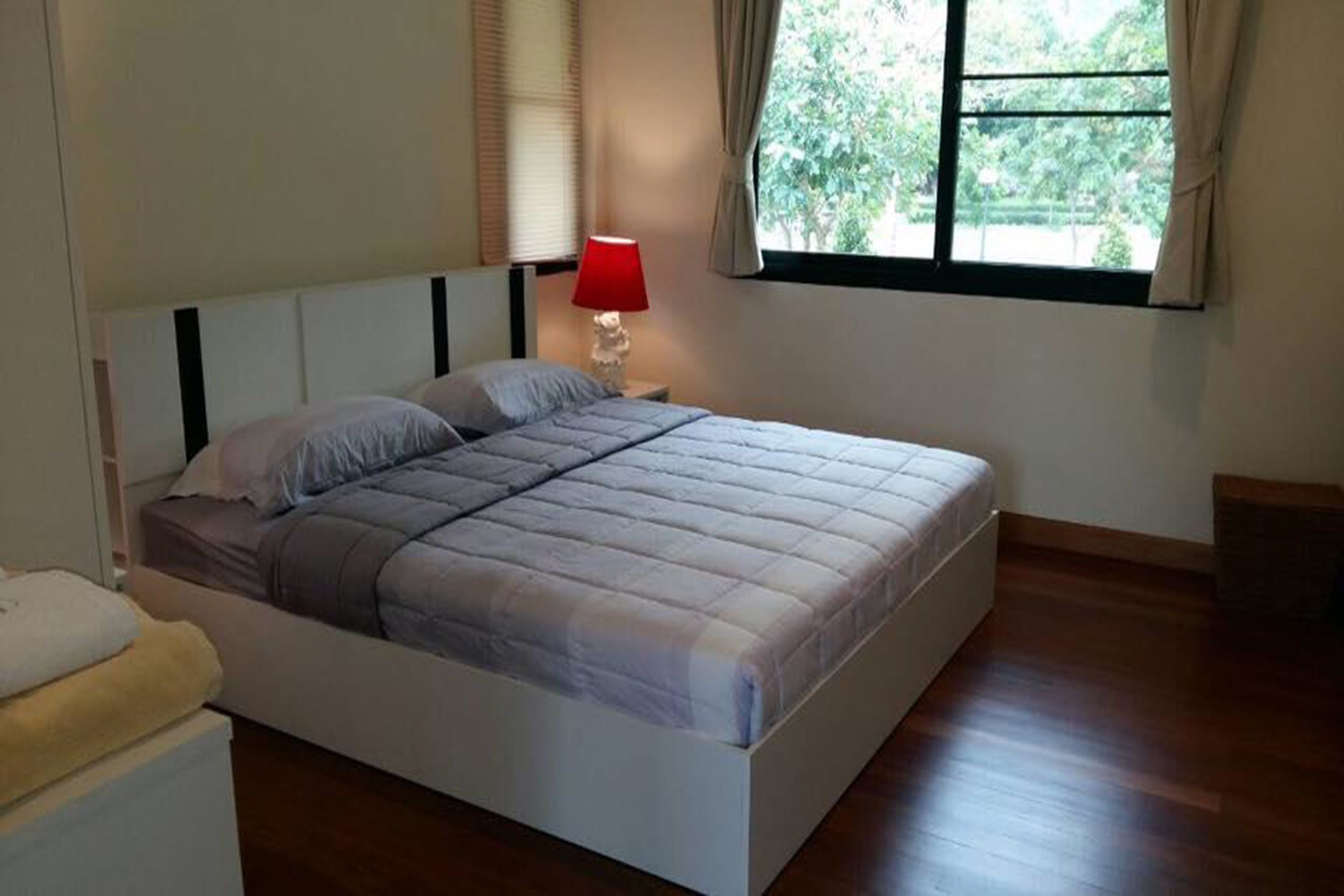 A bedroom with a bed, a nightstand with a lamp, a window with curtains, and a view of greenery outside.