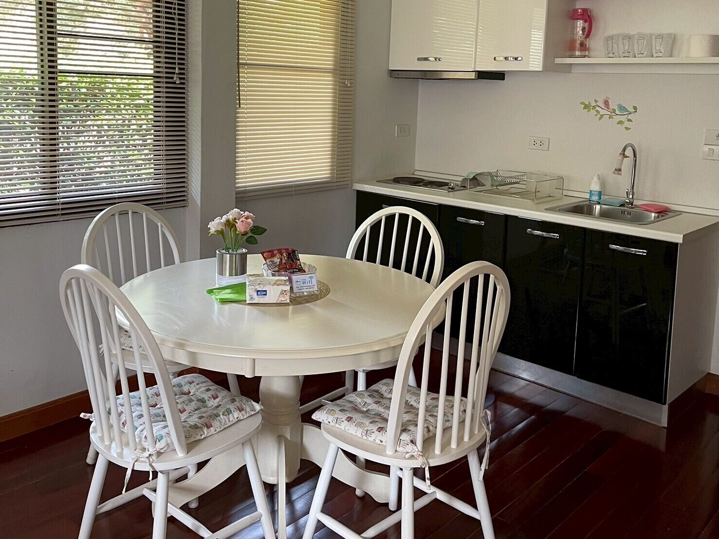 A dining area with a white round table and four white chairs, a vase with flowers, and a small dish with condiments.