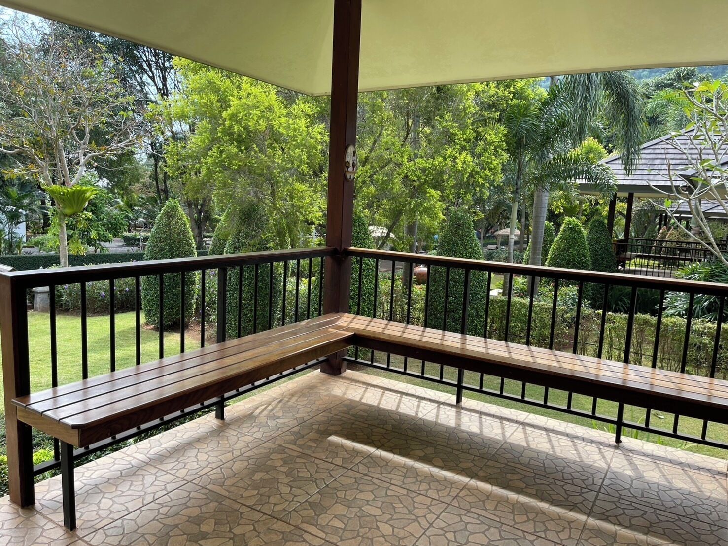 Un espacio de estar al aire libre cubierto con un banco, rodeado de un jardín con diversas árboles y arbustos.
