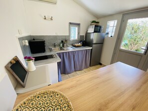 A compact kitchen with a microwave, toaster, and refrigerator. A small window provides natural light, and a table is positioned in the foreground.