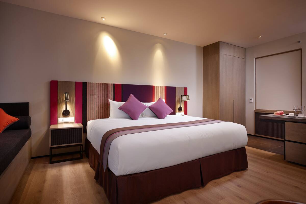 Ein modernes Hotelzimmer mit einem großen Bett, einem Nachttisch mit Lampe, einem kleinen Schreibtisch mit Stuhl und einem Kleiderschrank.