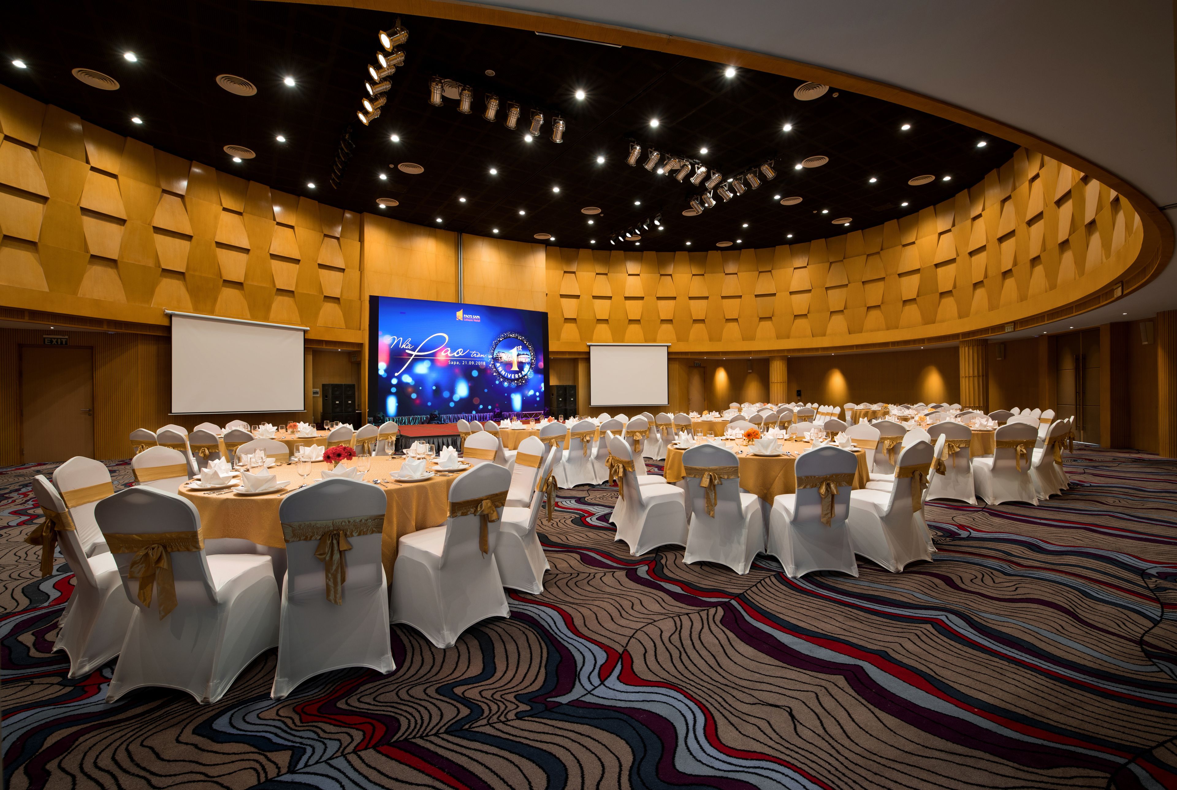 A conference room with round tables and white chairs, a large screen, and a stage.